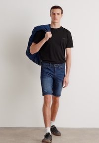T-shirt nera, pantaloni corti di denim blu e una giacca di denim appoggiata su una spalla. Modella con sneakers e calze bianche. Design semplice.