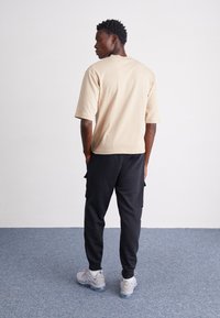 Beżowa koszulka z krótkim rękawem o prostym kroju, w połączeniu z czarnymi spodniami cargo i jasnoszarymi sneakersami, stojąca na jednolitym tle.