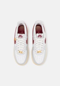White leather sneakers with maroon accents, perforated toe, thick laces, and gold hardware on the tongue. Rubber sole, classic silhouette.