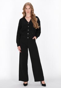 Czarny dzianinowy kardigan z srebrnymi zapięciami, posiadający dekolt w kształcie litery V i cekinową teksturę, zestawiony z szerokimi czarnymi spodniami.