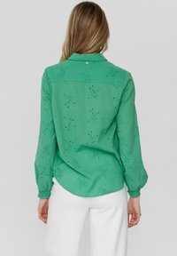 Camicia verde a maniche lunghe con tessuto testurizzato, ricami floreali e dettaglio con bottoni al colletto. Pantaloni bianchi sono parzialmente visibili.