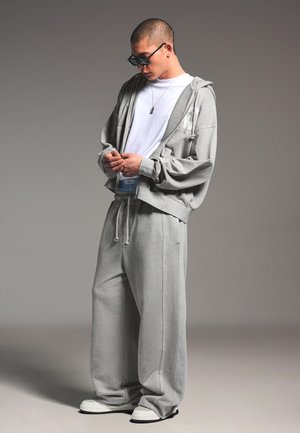 Bărbat tânăr purtând hanorac gri supradimensionat și pantaloni, cu adidași albi, privind în jos și ajustându-și hainele, pe un fundal gri simplu.