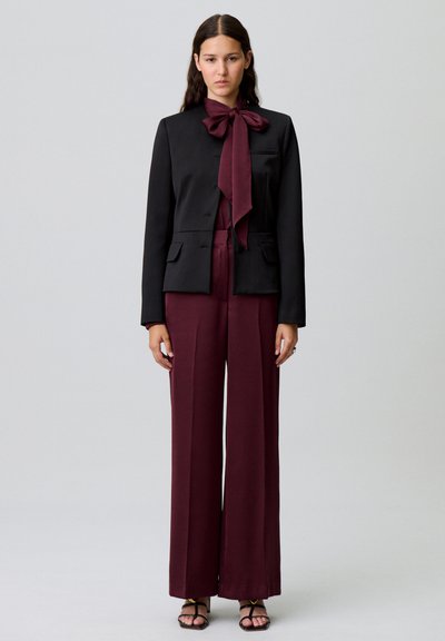 Blazer nero con un grande papillon bordeaux, abbinato a pantaloni bordeaux a gamba larga. Tessuto liscio con vestibilità su misura e dettagli con tasche.