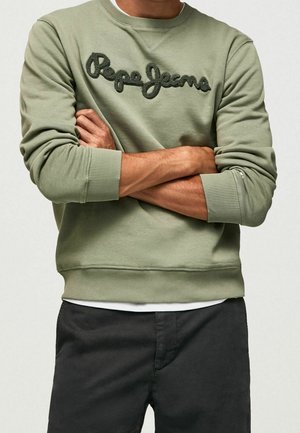 Mężczyzna w jasno oliwkowej bluzie z czarnym napisem "Pepe Jeans", ze skrzyżowanymi rękami, w połączeniu z czarnymi spodniami i widoczną białą podkoszulką.
