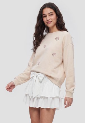 Ung kvinde med langt mørkt hår bærer beige sweatshirt med små dyreansigter og hvid lagdelt nederdel med stor sløjfe ved taljen.