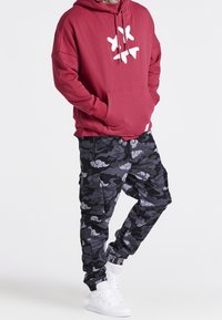 Felpa maroon con dettagli grafici bianchi, abbinata a pantaloni jogger camouflage neri e grigi con polsini elastici e tasche laterali. Sneakers bianche.