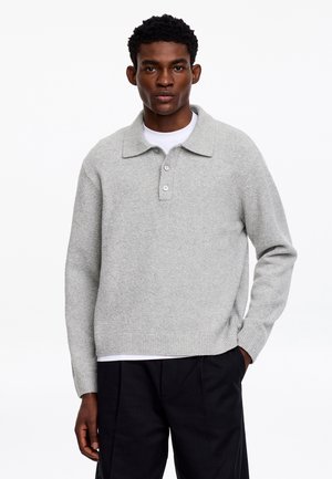 Mand iført en lysegrå strikket pullover med krave og knapper, hvid undertrøje og sorte bukser, stående med den ene hånd i lommen.