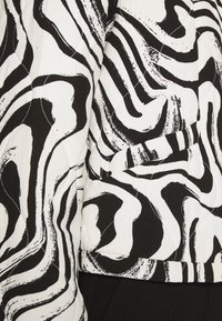 Motif tourbillonnant abstrait noir et blanc sur une veste en tissu matelassé avec une poche latérale et un pantalon noir visible.