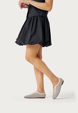 Mujer con una falda burbuja negra y zapatos planos blancos con cordones y patrones perforados en negro, de pie sobre una pierna contra un fondo liso.