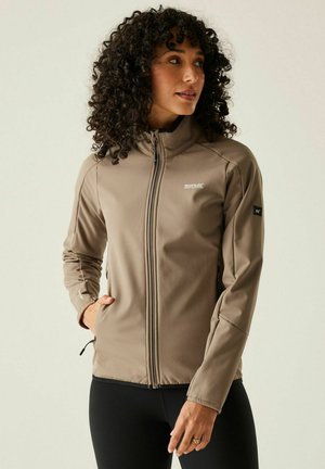 Femme aux cheveux bouclés portant une veste beige zippée avec les mains dans les poches, regardant sur le côté devant un fond clair uni.