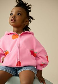 Rosa Zip-Up-Hoodie mit orangefarbenen Obstgrafiken, weichem Stoff und gerippten Bündchen, kombiniert mit Jeansshorts.