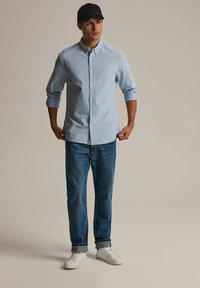 Jasnoniebieska koszula zapinana na guziki z długim rękawem, w zestawie z niebieskimi dżinsami i białymi trampkami. Koszula ma dyskretną logotyp na piersi.