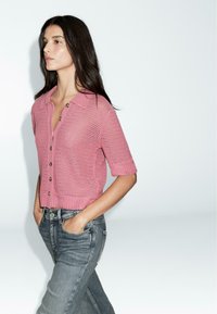 Rosa gestrickte kurzärmlige Strickjacke mit Kragen und Knopfverschluss, kombiniert mit blauen Jeans aus Denim. Sichtbares strukturiertes Webdetail.