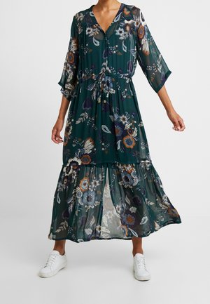 Femme portant une longue robe fleurie verte à manches 3/4 et des baskets blanches, debout avec les bras légèrement écartés.