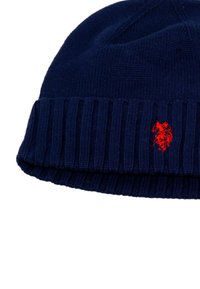 Căciulă tricotată de culoare bleumarin cu un model canalat la manșetă, având un mic emblema roșie pe față. Textură moale și vârf rotunjit.