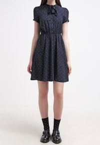 Marinblå klänning med korta ärmar och fluga, med vita prickar, en figursydd midja och en utsvängd kjol. Svart mönstrade strumpor och skor.