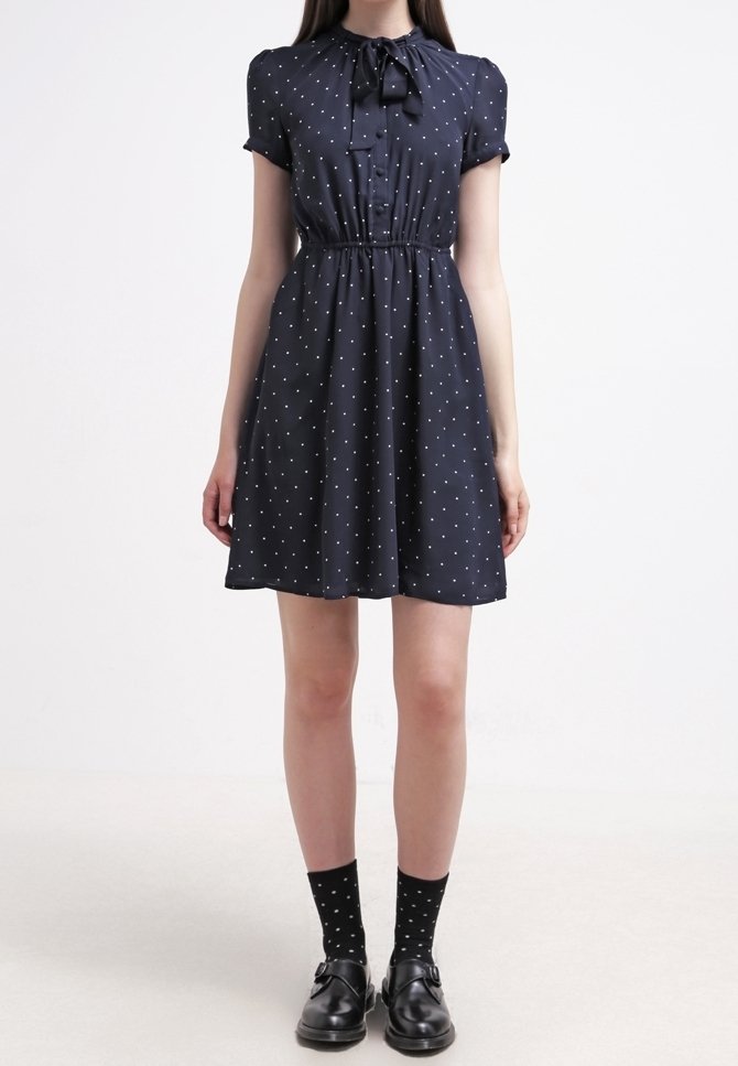 Marinblå klänning med korta ärmar och fluga, med vita prickar, en figursydd midja och en utsvängd kjol. Svart mönstrade strumpor och skor.