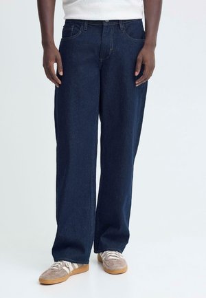Person, die dunkelblaue Jeans mit weitem Bein, ein weißes Top und beige Sneakers mit weißen Schnürsenkeln trägt, steht vor einem einfarbigen Hintergrund.