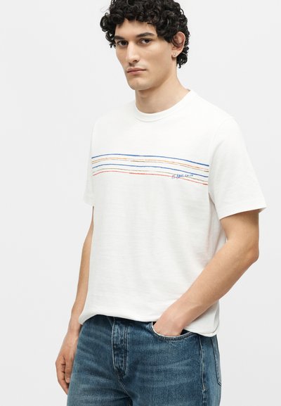 Νέος άνδρας με σγουρά μαλλιά που φοράει λευκό T-shirt με πολύχρωμες οριζόντιες ρίγες και μπλε τζιν, χέρι στην τσέπη, ουδέτερη έκφραση.