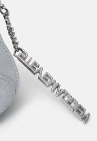 Grijze faux-suède tas met een metalen ketting en een hanger met het woord "WELKOM" in een strakke lettertype, met een gladde textuur.
