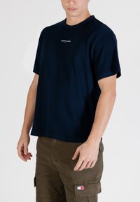 Maglietta in cotone blu navy con maniche corte e scollo rotondo, con un piccolo logo bianco sul petto, indossata con bermuda cargo color khaki.