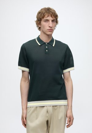 Jeune homme aux cheveux blonds bouclés, portant un polo vert foncé à manches courtes avec un liseré clair et un pantalon beige, debout devant un fond blanc uni.