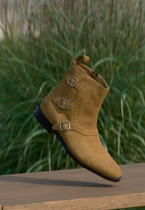 Beiger Wildleder-Knöchelstiefel mit drei silbernen Schnallen und dekorativen goldenen Nieten, ruhend auf einer Holzoberfläche mit Hintergrund aus hohem grünem Gras.