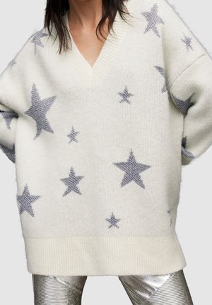 Osoba oblečená v nadměrně velkém bílém pleteném svetru se stříbrnými hvězdami a kovově stříbrných kalhotách, zobrazená od krku po střed stehen.