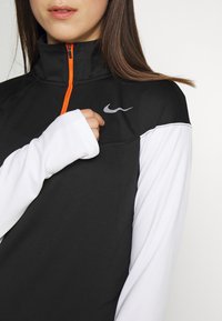 Mustapukuinen urheilutakki, jossa on neljänneksen vetoketju ja oranssi veto. Pitkät valkoiset hihat ja musta olkalevy, jossa on pieni valkoinen logo.