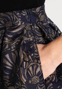 Květinová sukně v námořnické modři s metalickými zlatými detaily. Obsahuje texturovaný vzor, sklady a boční kapsy. Zapínání na zip vzadu.