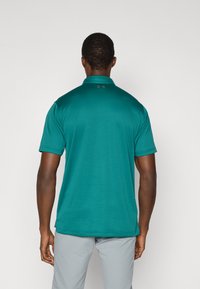 Τυρκουάζ πόλο μπλούζα με υφή υφάσματος, κοντά μανίκια και γιακά. Διαθέτει λογότυπο στην πλάτη, φορεμένη με ανοιχτό γκρι παντελόνι.