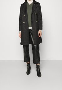 Cappotto trench doppiopetto nero con bottoni dorati, sovrapposto a un maglione verde e pantaloni cropped in finta pelle nera, abbinato a stivali neri alla caviglia.