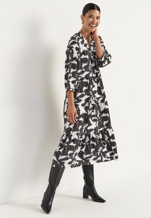 Langes, gestuftes Kleid aus weißem Stoff mit schwarzem abstraktem Blattmuster. Verfügt über 3/4 Ärmel und einen runden Ausschnitt. Trägt kniehohe schwarze Stiefel.