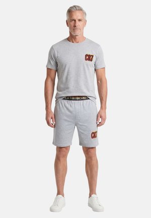 Uomo di mezza età con indosso una maglietta grigio chiaro e pantaloncini con loghi CR7 rossi e gialli, in piedi davanti a uno sfondo bianco uniforme.