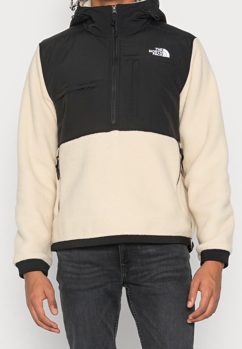 Mann in einer schwarzen und beigen Fleecejacke von The North Face mit Reißverschlusstaschen und schwarzen Jeans, stehend mit entspannten Armen.