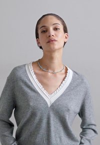 Γκρι πουλόβερ με λευκή ριγέ καπιτονέ λαιμόκοψη, μακριά μανίκια, απαλό υλικό, συνδυασμένο με ένα πολύχρωμο κολιέ από χάντρες.