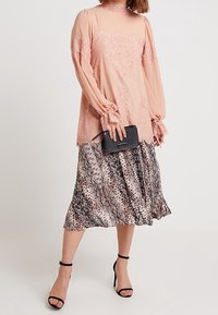 Långärmad topp i blush med spetsdetaljer, matchad med en mönstrad, flödande kjol i nyanser av rosa och svart. Accessoarer inkluderar en liten svart kuvertväska.