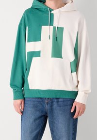 Zelená a bílá mikina s kapucí s velkým geometrickým vzorem, s přední kapsou a žebrovanými manžetami a lemem. Měkký textilní materiál.