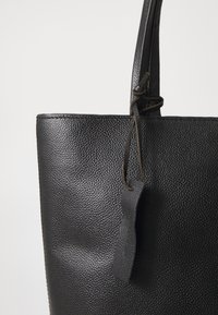 Borsa tote in pelle nera con superficie testurizzata, dotata di un'etichetta in pelle a forma di anello e spallacci sottili. Design semplice e strutturato.