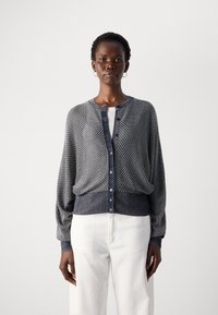 Strikket cardigan i marineblått og hvitt med et teksturert mønster, rund hals, knapper foran og ribbede mansjetter og kant. Brukt over en hvit topp.