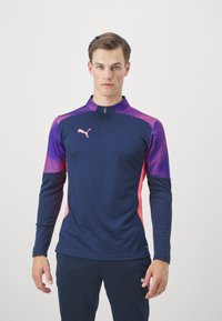 Marineblaue, sportliche Langarmoberteil mit einem Viertelreißverschluss, ausgestattet mit lila und pinken Musterakzenten auf den Schultern und Ärmeln. Puma-Logo angezeigt.