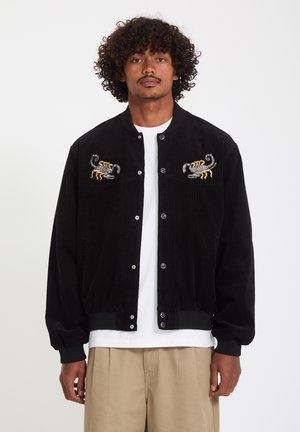 Homme aux cheveux bouclés portant une veste noire boutonnée avec des scorpions brodés sur la poitrine, une chemise blanche et un pantalon beige sur un fond uni.