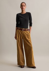 Vrouw staat in een zwarte langsliggende shirt, brede mosterdkleurige corduroybroek, bruine riem en bruine laarzen tegen een neutrale achtergrond.