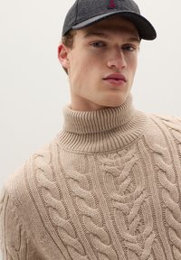 Maglione beige a collo alto con lavorazione a trecce e motivi testurizzati; indossato con un berretto scuro con logo rosso. Vista ravvicinata del capo e degli accessori.