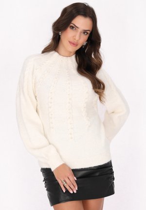 Maglione bianco lavorato a maglia con un motivo testurizzato e maniche a palloncino, abbinato a una mini gonna di pelle nera. Accentuato con piccoli dettagli decorativi.