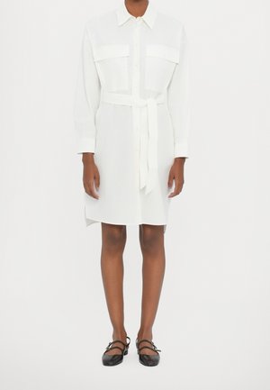 Femme portant une robe chemise blanche à manches longues ceinturée avec deux poches poitrine et des chaussures plates noires avec brides à la cheville.