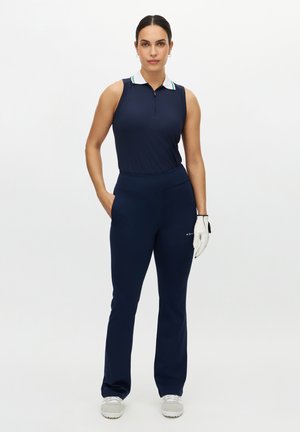 Marineblaues ärmelloses Polo mit Reißverschluss am Kragen und weißem Kragenstreifen, kombiniert mit marineblauen, eng anliegenden Hosen und weißen Golfschuhen mit Handschuh.