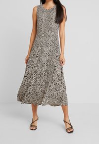 Hihaton midi-mekko vaaleanbeigessä, jossa on mustia pilkkuja, A-linjainen siluetti, valuva kangas, yhdistettynä mustiin hihnalasia sandaaleihin.