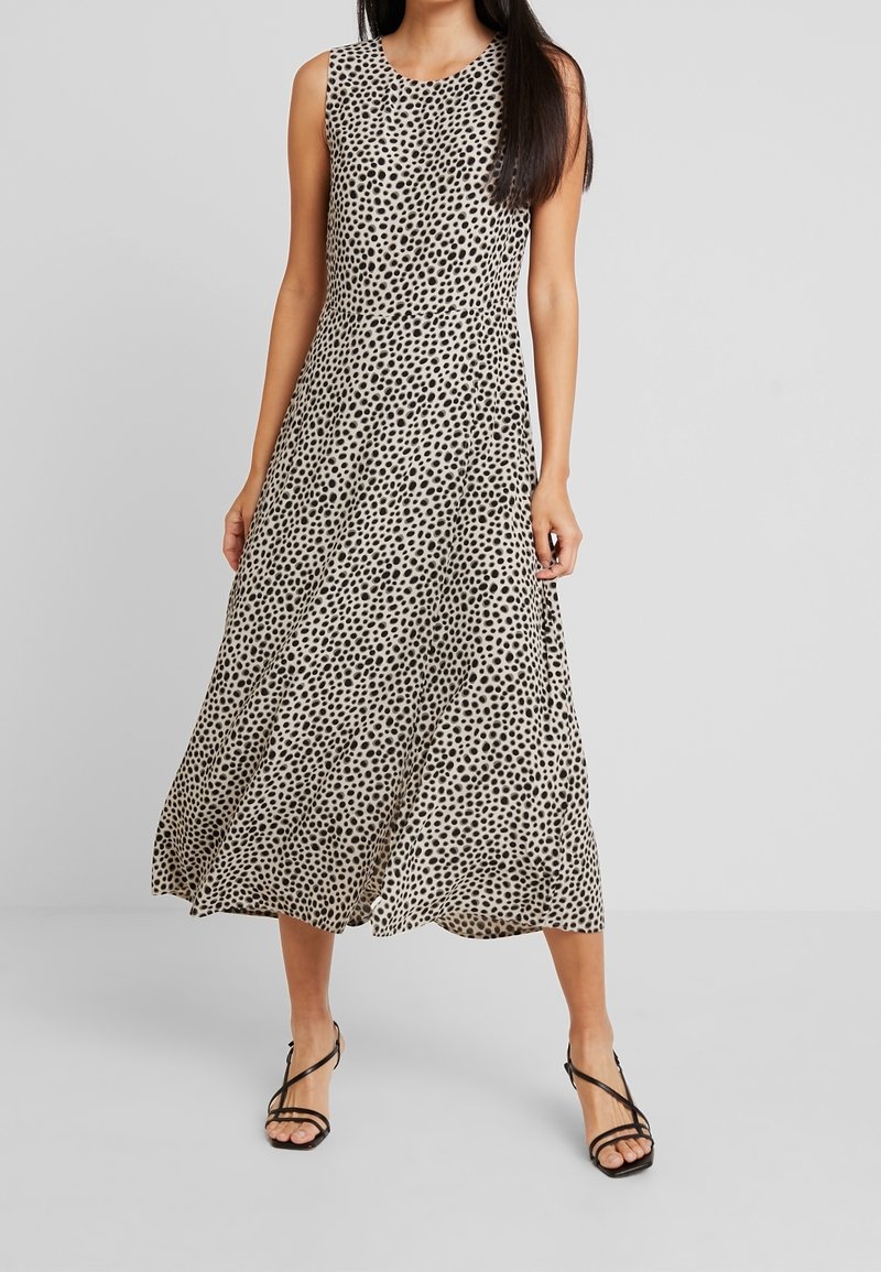 Hihaton midi-mekko vaaleanbeigessä, jossa on mustia pilkkuja, A-linjainen siluetti, valuva kangas, yhdistettynä mustiin hihnalasia sandaaleihin.