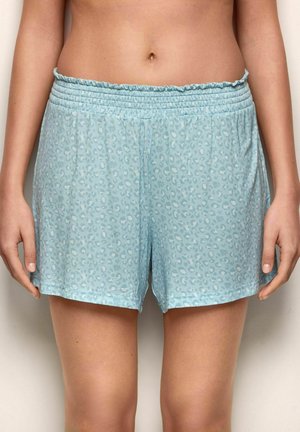 Donna che indossa pantaloncini elasticizzati in vita di colore azzurro chiaro con stampa leopardo sottile, in piedi davanti a uno sfondo neutro con le mani rilassate ai lati.